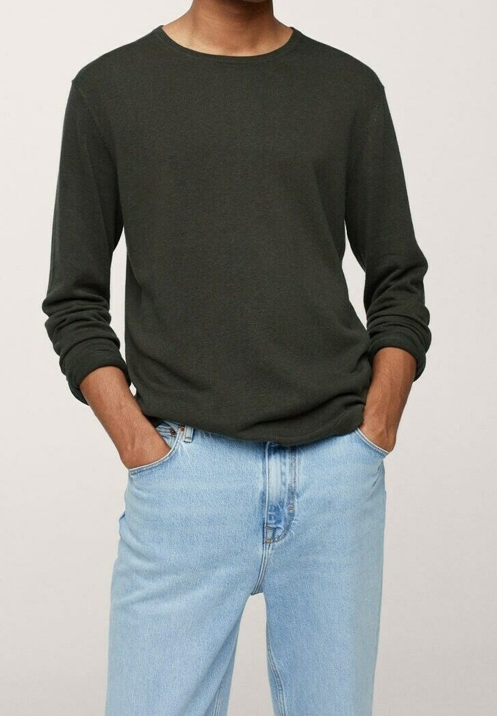 Homme portant un pull à manches longues vert foncé et un jean bleu clair, les mains posées négligemment dans les poches, sur un fond uni.