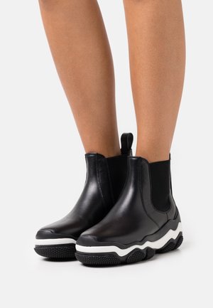 Legs wearing black leather ankle boots with thick white and black textured soles on a white background.
