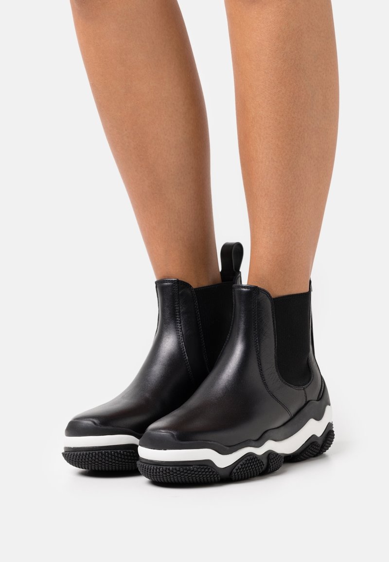 Piernas con botas de tobillo de cuero negro con suelas gruesas de texturas blancas y negras sobre un fondo blanco.