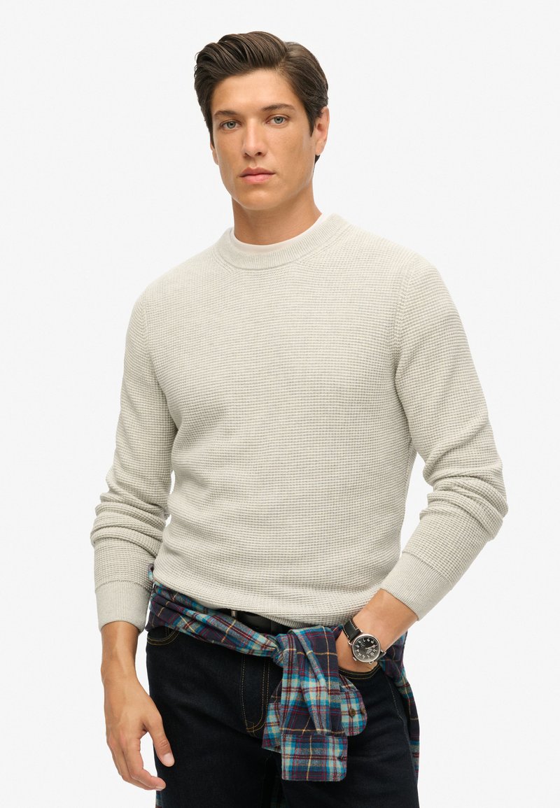 Lichtgrijze textuurtrui met een ronde hals, gecombineerd met donkere jeans en een blauwe geruite shirt om de taille geknoopt. Zwarte horloge om de pols.