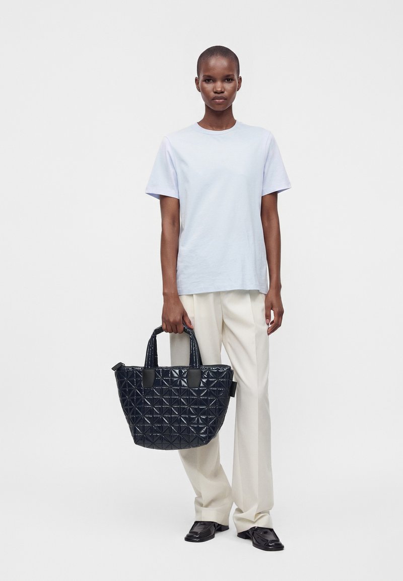 Une personne debout porte une chemise bleu clair à manches courtes, un pantalon beige, des chaussures noires et tient un grand sac à main noir matelassé dans sa main droite.