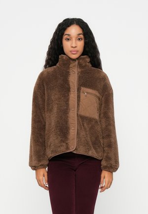 Veste en polaire marron avec col haut, fermeture à pression sur le devant et poche zippée. Tissu texturé avec une apparence douce et moelleuse.