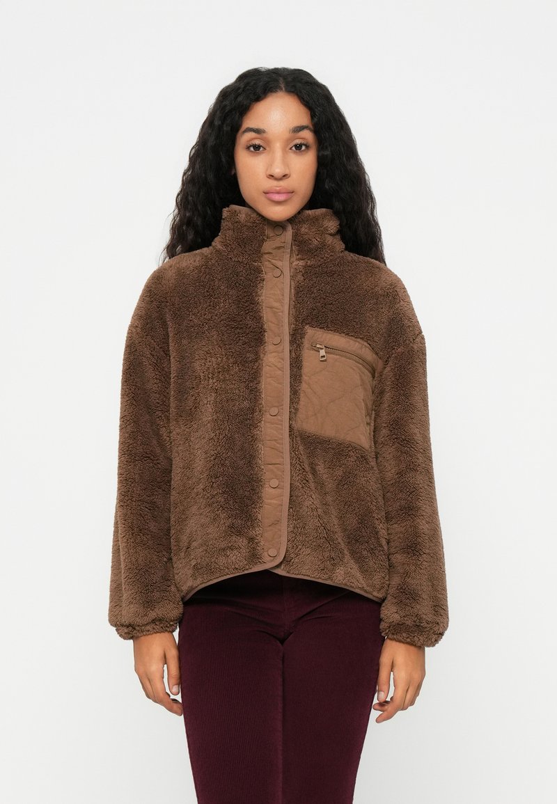 Veste en polaire marron avec col haut, fermeture à pression sur le devant et poche zippée. Tissu texturé avec une apparence douce et moelleuse.