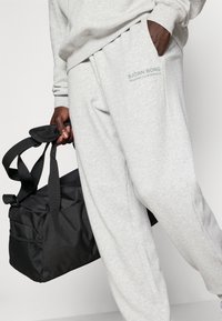 Pilkos sportinės kelnės su kišene, turinčios "BJÖRN BORG" prekės ženklą. Juodas sportinis krepšys su tekstūruota medžiaga ir tinklo panelė, laikomas rankoje.