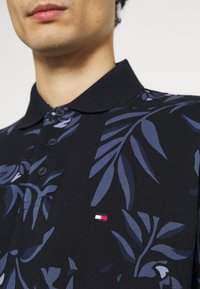 Homme portant un polo noir avec des motifs de feuilles bleues et un petit logo rectangulaire rouge, blanc et bleu marine sur la poitrine.