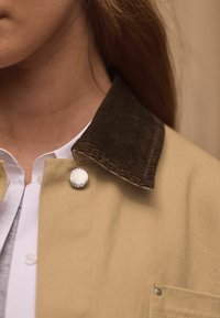 Beige pusero takki tummanruskealla corduroy-kauluksella, hopeisella nepparikinnityksellä ja kaulusreunassa näkyvällä tekstitjustuksella.