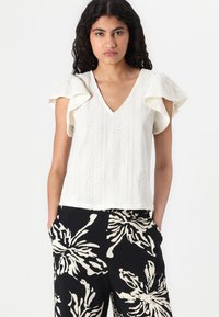 Bluză albă cu mânecă scurtă și detalii de dantelă, cu decolteu în V. Asortată cu pantaloni negri, care au un model floral mare alb.