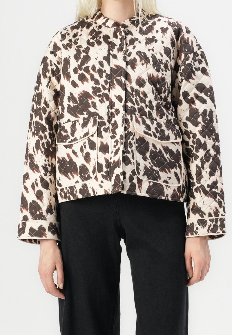 Femme portant une veste matelassée avec des taches abstraites marron et blanches et de grandes poches avant à rabat, associée à un pantalon noir.