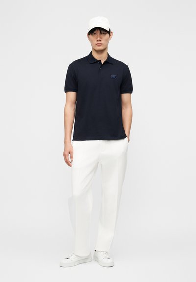 Άνδρας με λευκό καπέλο, μπλε σκούρο πόλο μπλούζα, λευκό παντελόνι και λευκά αθλητικά παπούτσια, στέκεται μπροστά σε μία απλή φωτεινή φόντο.