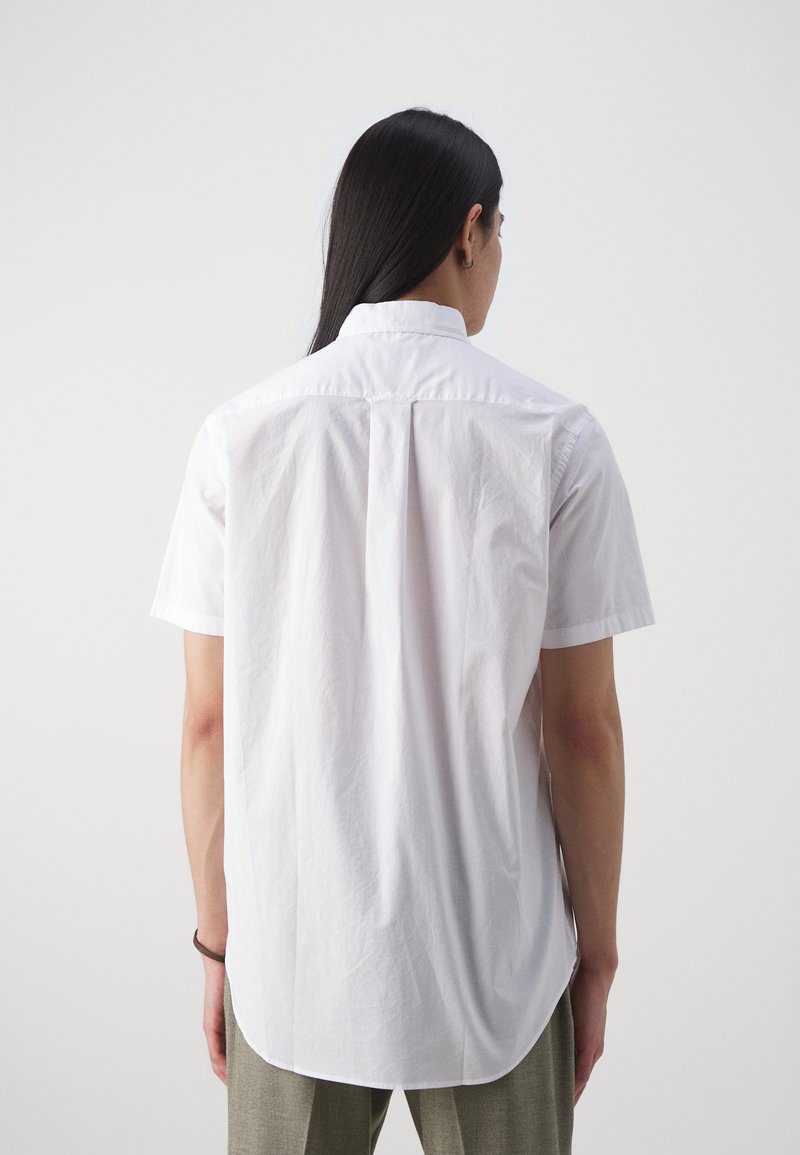 Chemise blanche à manches courtes, avec une texture lisse, un ourlet arrondi et un design de empiècement au dos, présentant une esthétique minimaliste.