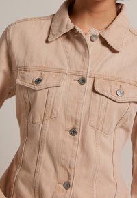 Ljusbeige denimjacka med en strukturerad design, utrustad med en spetsig krage, två bröstfickor och silverfärgade knappar.