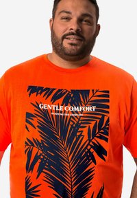 Krátký rukáv oranžové tričko s tmavě modrým potiskem palmového listu a bílým textem "JEMNÉ POHODLÍ UŽÍVÁNÍ DOBRÉHO ŽIVOTA" uprostřed.