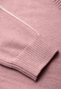 Rosa Pullover mit einer weichen Textur, ausgestattet mit gerippten Bündchen und einem kontrastierenden hellgrünen Streifen entlang der Naht. Nahaufnahme des Stoffes.