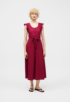 Kvinne med kort krøllete blondt hår som står, iført en ermeløs mørkerød midikjole med knyting i midjen og svarte flip-flops, nøytral bakgrunn.