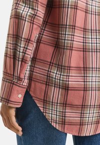 Close-up van een roze en bruine geruite shirtmanchet met een knoop, gedragen met blauwe jeans, die een zijuitsnede en een deel van de hand van een persoon toont.
