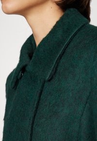 Gros plan d'une personne portant un manteau en laine duveteuse vert foncé avec un large col et des détails de boutons, montrant son cou et la ligne de sa mâchoire.
