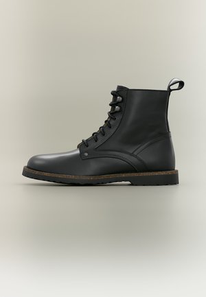 Bottine noire en cuir à lacets avec languette, bordure marron sur la semelle et semelle extérieure en caoutchouc robuste sur fond neutre.