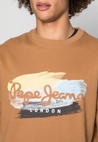 Primo piano di una persona che indossa un felpa marrone con il testo "Pepe Jeans London" su un design di pennellate gialle, bianche, azzurro chiaro e nere.