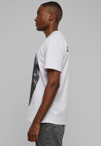 Valge puuvillane T-särk, mille küljel on suur musta ja halli graafikaga pilt, lühikeste varrukatega ja sirge äärega, kantud hallide teksadega.