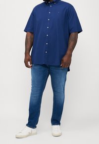 Man som bär en blå kortärmad skjorta, blekta blå jeans och vita sneakers står mot en neutral bakgrund.