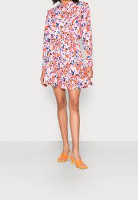 Robe imprimée florale avec manches longues, ourlet volanté et taille cintrée, dans des tons de rose, orange et bleu. Associée à des sandales à talons orange.