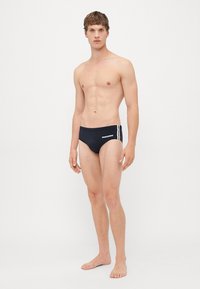 Junger Mann, der barfuß steht und schwarze Badehose mit weißen Seitenstreifen und dem "DSQUARED2"-Logo auf der linken Seite trägt.