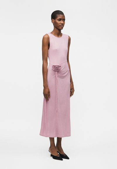 Γυναίκα φορούν χωρίς μανίκια, μακρύ φόρεμα σε σκότινο ροζ χρώμα με πιέτες και υφασμάτινο τριαντάφυλλο στη μέση, σε συνδυασμό με μαύρα παπούτσια με χαμηλό τακούνι.