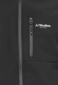 Black fleece jacket featuring a zippered chest pocket, reflective gray zippers, and a printed logo "JJ Studios. Jack & Jones" on the front.