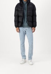 Uomo che indossa un giubbotto imbottito nero, un sudore con zip quarti blu, jeans azzurri e sneakers bianche, in piedi su uno sfondo semplice.