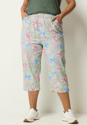 Underkrop iført lysegrønne korte bukser med pink og blå blomsterprint, hænder i lommerne, stylet med hvide sneakers.