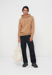 Felpa marrone con tasca frontale, caratterizzata da un piccolo logo giallo. Abbinata a pantaloni cargo blu navy e con una borsa tote beige. Sneakers nere.