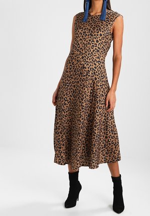 Žena v midi šatech bez rukávů s hnědým a černým leopardím vzorem, doplněná o černé kotníkové boty na podpatku a modré náušnice se střapci.