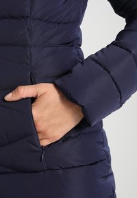 Marinblå puffjacka med quiltat mönster, försedda med en ficka med dragkedja. Ärmen är något rynkad vid handleden för en snug passform.