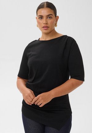 Femme aux cheveux foncés attachés, portant des boucles d’oreilles en or, un haut asymétrique noir à manches courtes et un pantalon sombre, debout devant un fond clair uni.