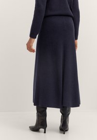 Person in einem langen, marineblauen Strickrock, passendem marineblauen Langarmoberteil und schwarzen Lederstiefeln mit mittelhohem Absatz, vor einem neutralen Hintergrund stehend.