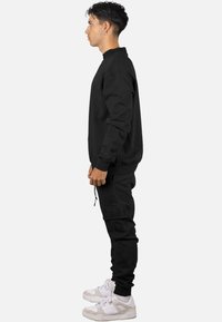 Sudadera negra con cuello alto y puños acanalados, combinada con pantalones joggers negros que tienen un parche texturizado con la palabra "DARK", usada con zapatillas blancas.