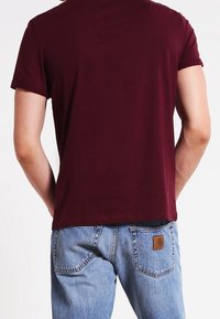 Camiseta de manga corta burdeos hecha de algodón suave, combinada con jeans de mezclilla de color azul claro que presentan una etiqueta de marca de cuero marrón en el bolsillo trasero.