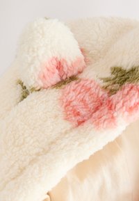 Gorro de forro polar color crema con patrones florales en rosa y verde y un pompón en la parte superior, descansando sobre un fondo de tela clara.