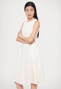 Λευκό αμάνικο φόρεμα με εφαρμοστό μπούστο και φαρδιά φούστα, κατασκευασμένο από λείο ύφασμα. Απλός σχεδιασμός, μέχρι το γόνατο, χωρίς ορατά σχέδια ή διακοσμητικά στοιχεία.