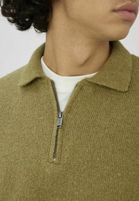 Pull-over tricot vert olive avec un col et une fermeture à glissière à mi-hauteur. Tissu texturé d'apparence douce, porté par-dessus une chemise blanche.