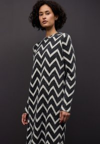 Šaty se svetrovým vzorem chevron v černé a bílé barvě, s volným střihem, dlouhými rukávy a měkkou, strukturovanou tkaninou.