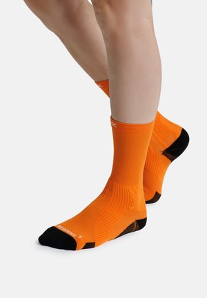 Benens met feloranje sportsokken met zwarte hielen en tenen, staand op een witte ondergrond.