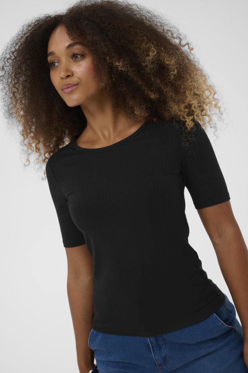 Femeie cu păr ondulat voluminos, îmbrăcată într-un tricou scurt, negru, cu neoptoare și blugi, pe un fundal simplu.