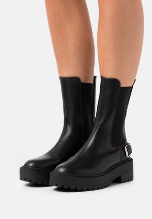 Botines de cuero negro con paneles laterales elásticos, suela de goma gruesa y un acento decorativo de hebilla en el lado exterior. Tacón plano, textura suave.