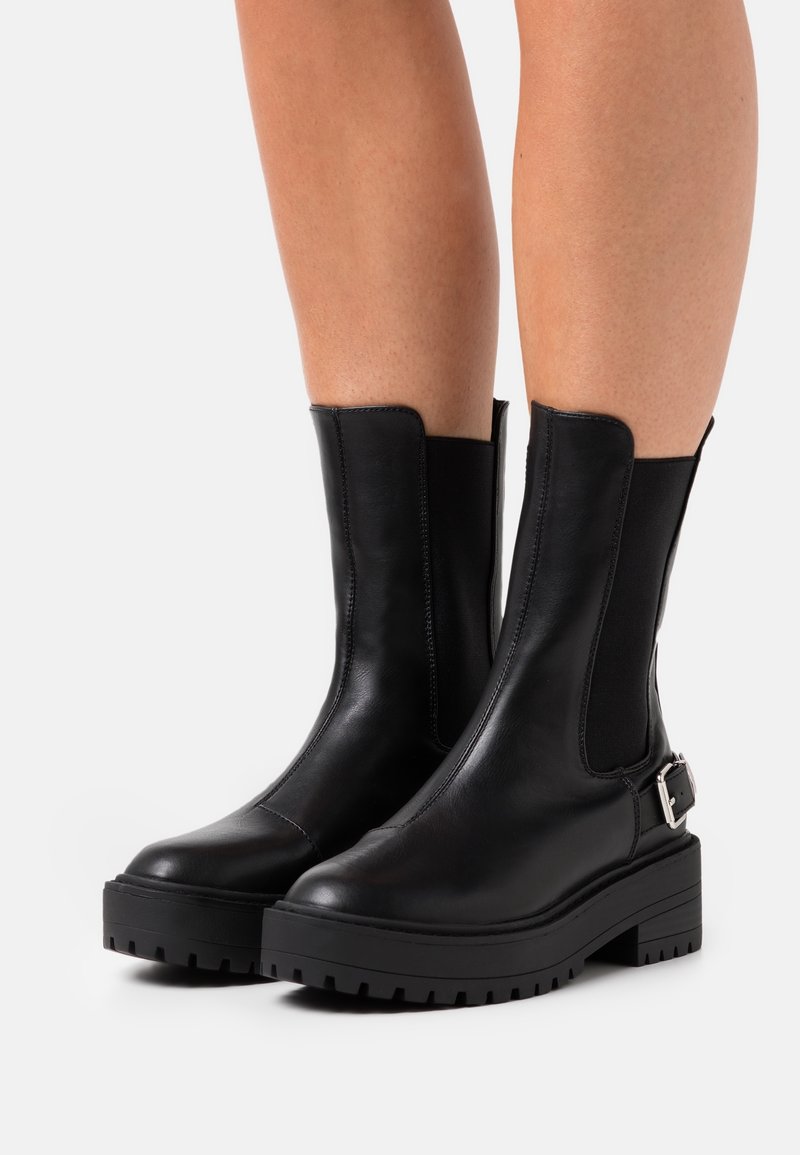 Botines de cuero negro con paneles laterales elásticos, suela de goma gruesa y un acento decorativo de hebilla en el lado exterior. Tacón plano, textura suave.