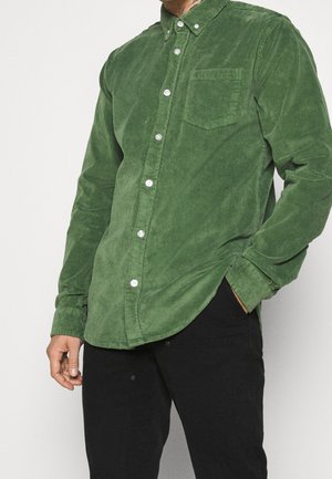 Mann trägt ein langärmeliges grünes Cord-Hemd mit Brusttasche und schwarzen Hosen, eine Hand in der Tasche.