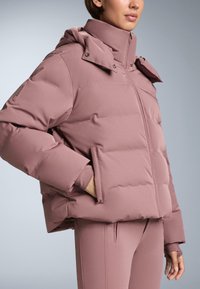 Ružičasta pernata jakna s visokim ovratnikom, patentnim zatvaračem sprijeda i kopčama na pritisak. Ima elastične manžete i bočne džepove, izrađena je od glatkog materijala.