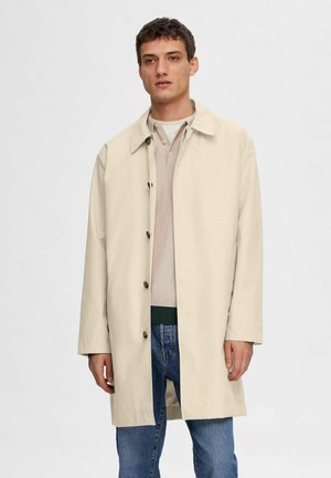Cappotto beige, a lunghezza ginocchio con colletto e chiusura a bottoni, realizzato in materiale liscio, abbinato a un polo chiaro e jeans in denim.