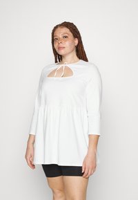 Blusa branca de manga comprida com bainha franzida, apresentando um recorte em forma de olho na gola amarrado com uma fita. Tecido suave e corte solto.