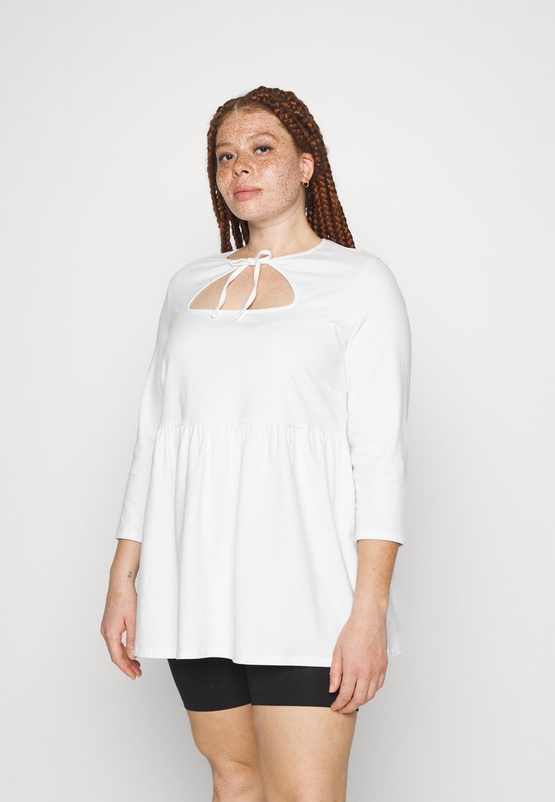 Blusa branca de manga comprida com bainha franzida, apresentando um recorte em forma de olho na gola amarrado com uma fita. Tecido suave e corte solto.